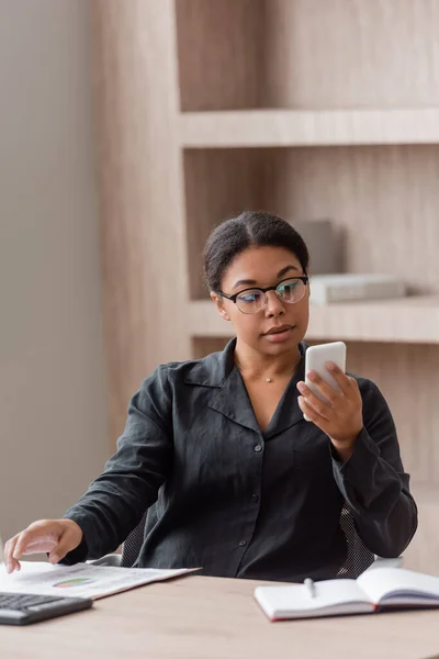 Brunette multiracial manager in eyeglasses looking at mobile phone while looking at smartphone near blurred notebook — Photo de stock