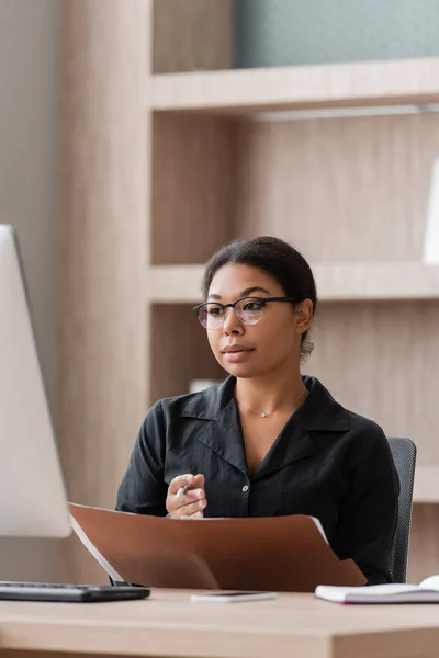Serious multiracial businesswoman in eyeglasses holding folder and looking at blurred computer monitor — Fotografia de Stock