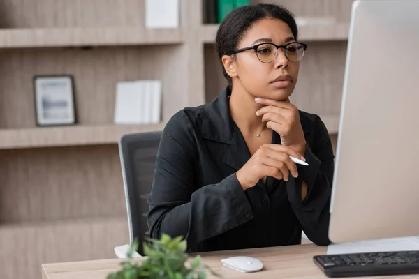 Thoughtful multiracial manager in eyeglasses holding pen and looking at computer monitor in office — Foto stock