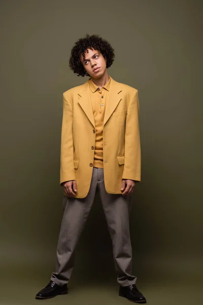 Full length of young african american guy in yellow jacket and pants standing on olive grey background — Stock Photo
