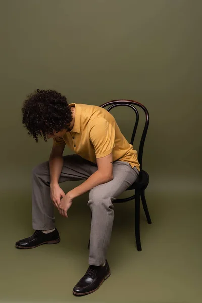 Full length of african american man in yellow polo shirt and black leather shoes sitting on chair with bowed head on grey green background - foto de stock