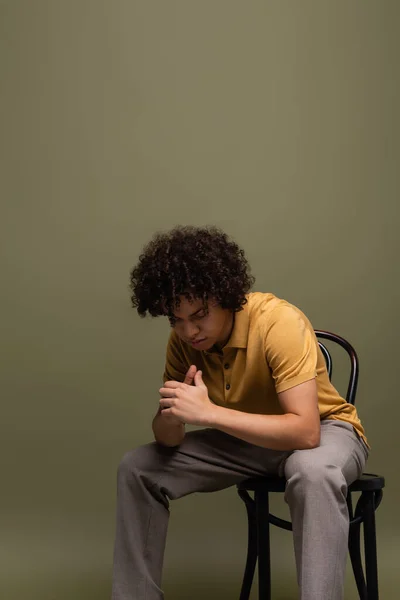 Curly african american man in yellow polo shirt and trousers sitting on chair with bowed head on grey green background — Stock Photo