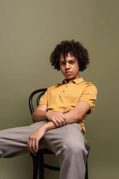 Trendy african american man in yellow polo shirt sitting on chair and looking at camera on grey background — Fotografia de Stock
