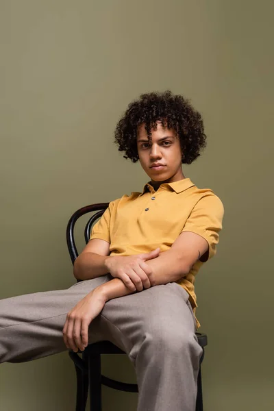 Trendy african american man in yellow polo shirt sitting on chair and looking at camera on grey background — Stock Photo