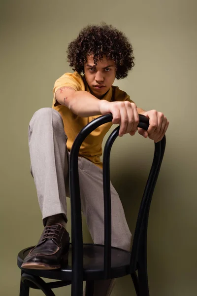 Young african american man in trendy casual attire stepping on chair and looking at camera on grey green background — Stock Photo