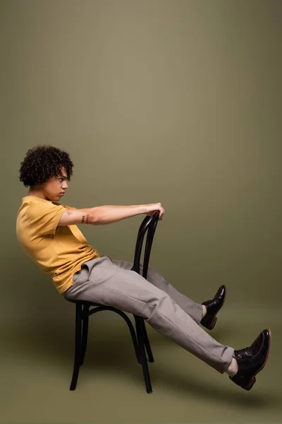 Vue latérale de l'homme afro-américain tatoué en t-shirt jaune et chaussures en cuir noir posant sur chaise sur fond gris-vert — Photo de stock