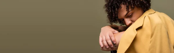 African american guy with curly hair obscuring face with yellow blazer while sitting with crossed arms isolated on grey, banner — Stock Photo