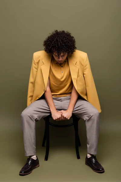 Vue de face de l'homme afro-américain en veste jaune et chaussures en cuir noir assis sur une chaise avec tête inclinée sur fond gris-vert — Photo de stock
