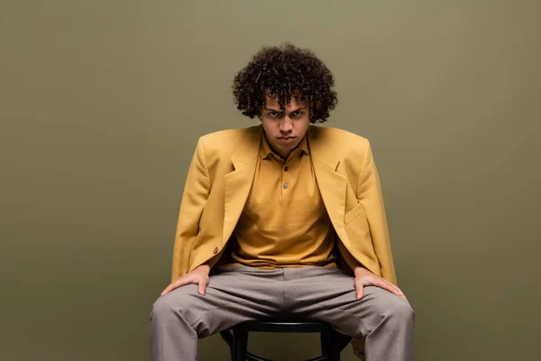 Front view of fashionable african american man in yellow blazer and trousers sitting and looking at camera on grey background — Stock Photo