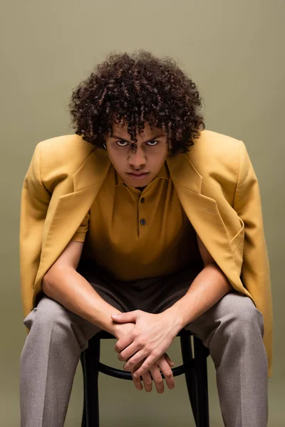 Young and confident african american man in yellow jacket looking at camera while sitting isolated on grey — Stock Photo