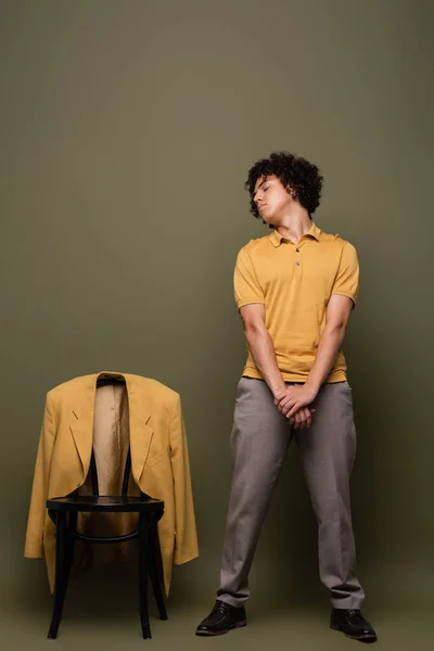 Homme afro-américain de toute la longueur en polo jaune et pantalon debout avec les yeux fermés près de la chaise avec blazer sur fond gris-vert — Photo de stock