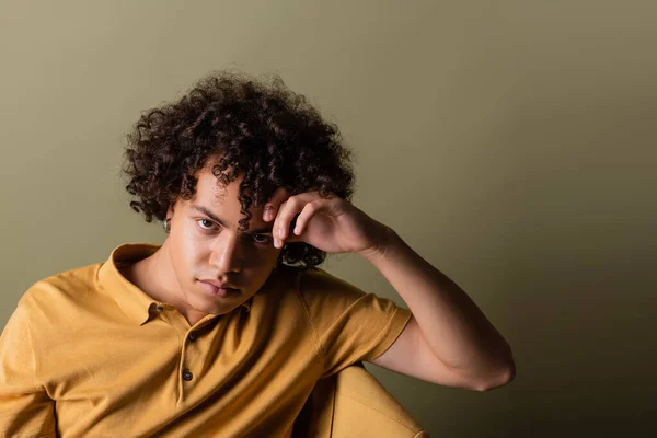 Curly african american man in yellow polo shirt holding hand near head and looking at camera while sitting on grey green background — Stock Photo