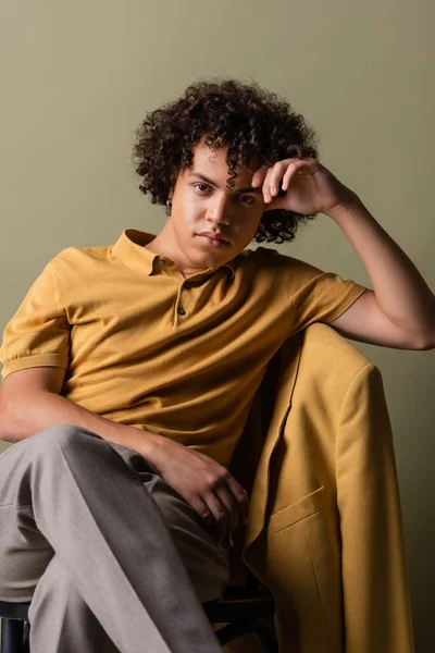Trendy african american man in yellow polo shirt sitting with crossed legs and hand near head on grey background — Stock Photo