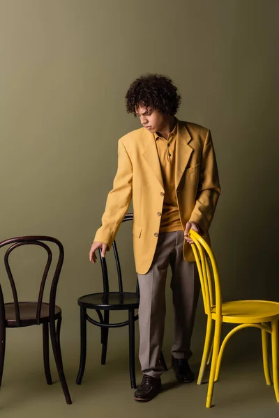 Piena lunghezza dell'uomo afro-americano in abiti casual alla moda in piedi vicino a sedie nere e gialle su sfondo grigio oliva — Foto stock