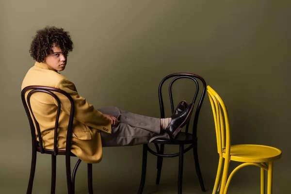 Повна довжина стильного афроамериканця в жовтому блістері, що сидить на чорних стільцях на сіро-зеленому фоні — стокове фото