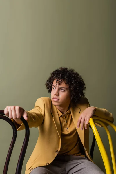 Hombre afroamericano con estilo en chaqueta amarilla mirando hacia otro lado cerca de sillas negras y amarillas aisladas en gris - foto de stock