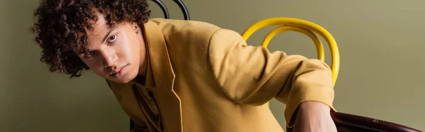 Афроамериканський чоловік з кучерявим брюнетка волоссям позує в жовтій куртці біля стільців на сірому фоні, банер — стокове фото
