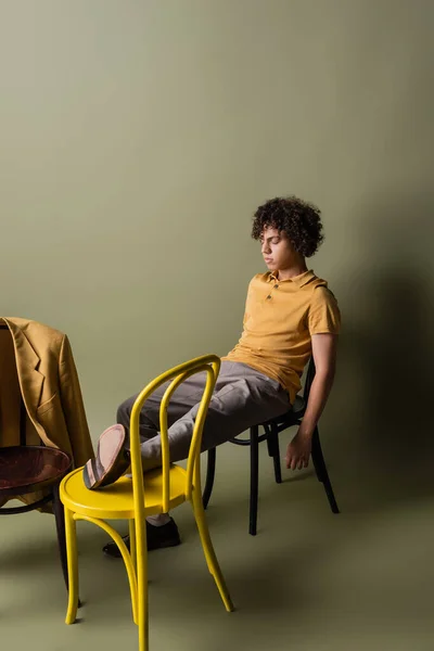 Full length of african american man with closed eyes sitting on black and yellow chairs near trendy blazer on grey green background — Foto stock