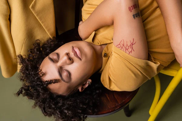 Top view of tattooed african american man in yellow polo shirt lying with closed eyes on grey green background — Foto stock