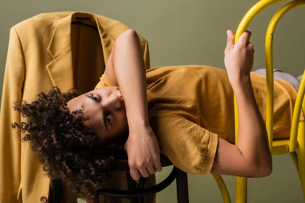 Молодий афроамериканський чоловік дивиться на камеру, лежачи на стільцях біля модного жовтого блейзера ізольовані на сірому — стокове фото