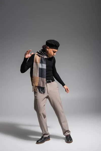 Full length of stylish african american man in black sweater and plaid scarf with beret posing on grey background - foto de stock