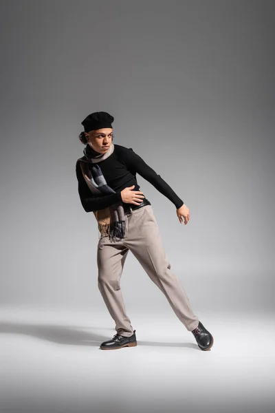 Full length of african american man in stylish beret and black sweater with checkered scarf looking away while posing on grey background — Fotografia de Stock