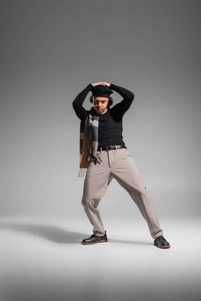 Comprimento total de homem americano africano na moda em boina preta e pulôver com cachecol posando com as mãos acima da cabeça sobre fundo cinza — Fotografia de Stock