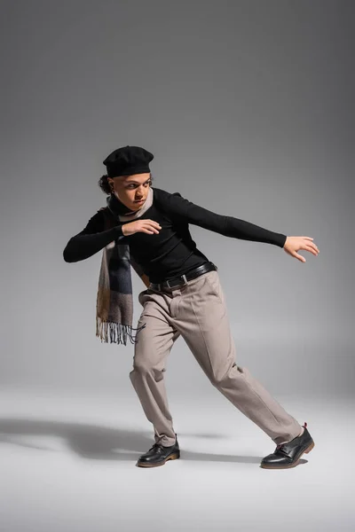 Full length of african american man in black beret and sweater with plaid scarf looking away on grey background — Stock Photo