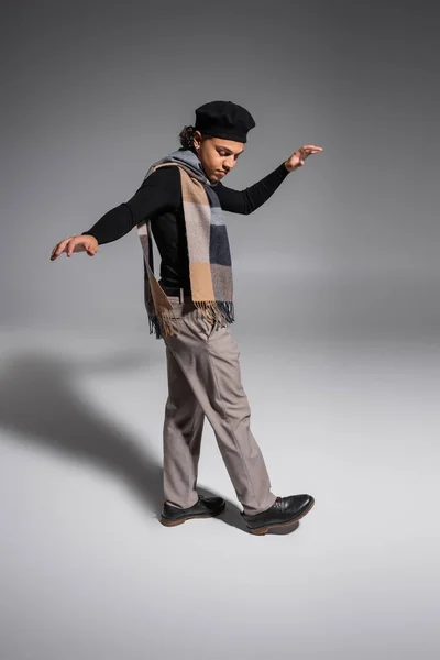 По всей длине африканский американец в черном свитере с беретом и клетчатым шарфом, шагающий на сером фоне — стоковое фото