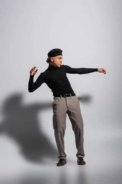 Full length of fashionable african american guy in black beret and turtleneck looking away while standing on grey background - foto de stock