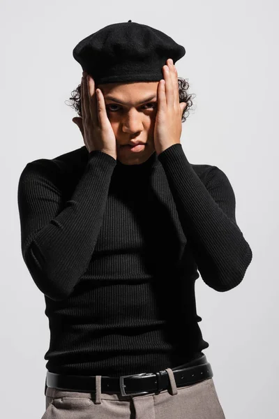 Stylish african american man in black turtleneck and beret holding hands near face and looking at camera isolated on grey — Stock Photo