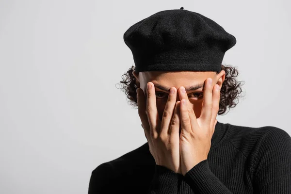 Jeune homme afro-américain en noir élégant béret obscurcissant le visage avec les mains et regardant la caméra isolée sur gris — Photo de stock