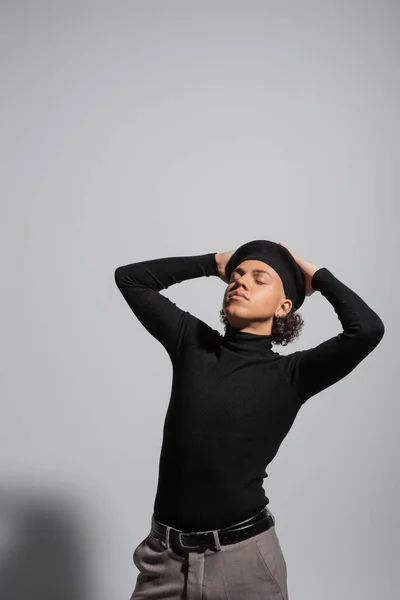 African american guy in black pullover and beret posing with closed eyes and hands on head on grey background — Stock Photo