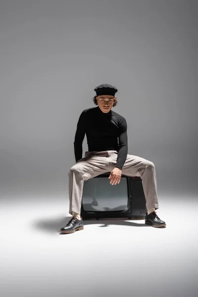 Full length of trendy african american man in black turtleneck and pants with leather shoes sitting on vintage tv set on grey background — Stock Photo