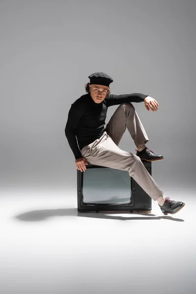 Full length of african american man in stylish beret and black pullover sitting on retro tv and looking at camera on grey background — Stock Photo