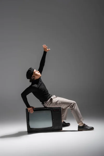 Side view of african american man in fashionable casual attire sitting on vintage tv set with raised hand on grey background — Stock Photo