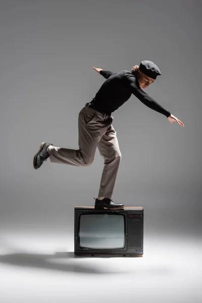 Full length of trendy african american man in black sweater and trousers balancing on vintage tv set on grey background — Stock Photo