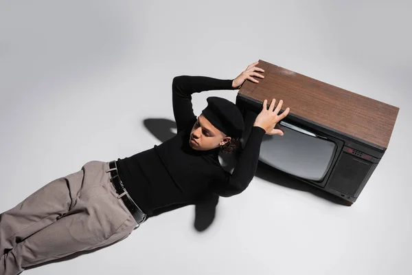 Top view of stylish african american man in black beret and turtleneck lying with closed eyes near retro tv set on grey background — Stock Photo