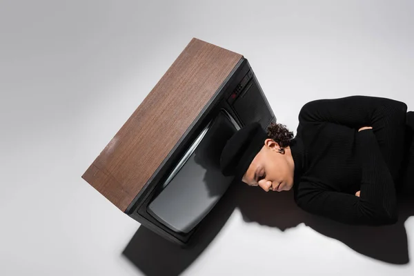Top view of african american man in black pullover and beret lying with crossed arms near retro tv set on grey background — Stock Photo