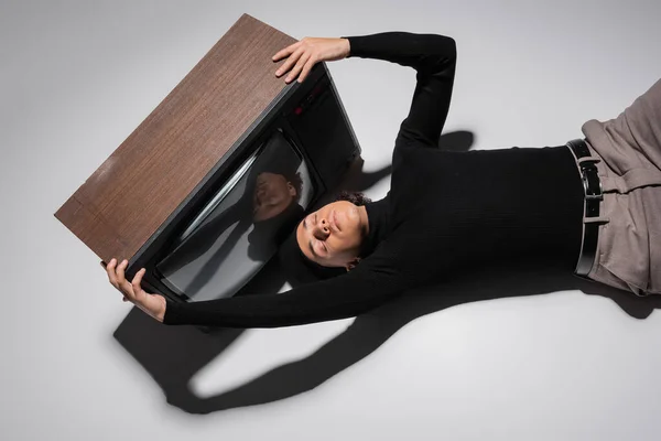 Top view of fashionable african american man in black turtleneck and trousers lying near vintage tv set on grey background — Stock Photo