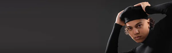 Jovem homem americano africano em camisola preta ajustando boina elegante enquanto olha para a câmera isolada em cinza escuro, banner — Fotografia de Stock