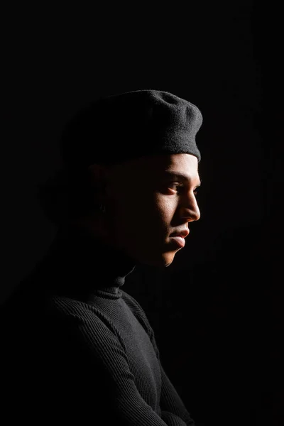 Profile of african american man with shaded face wearing dark beret and sweater isolated on black — Stockfoto