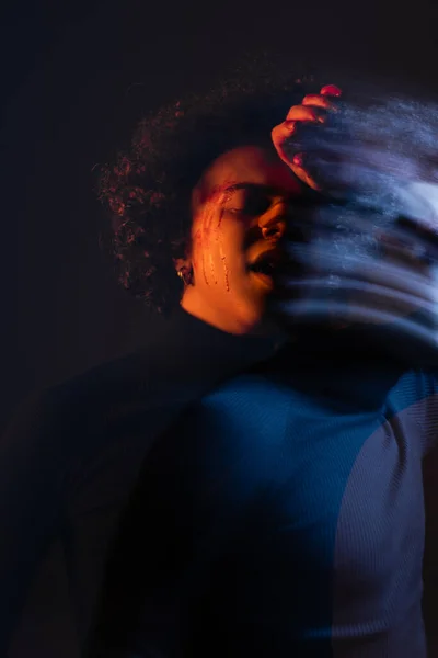 Double exposure of wounded african american man standing with hand near bleeding face on dark background with red and blue light - foto de stock