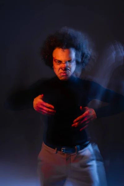 Desenfoque de movimiento del hombre afroamericano agresivo con trastorno bipolar y cara ensangrentada mirando a la cámara en la oscuridad con luz naranja y azul - foto de stock