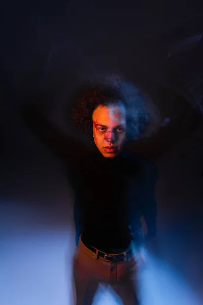 Injured african american man with bipolar disorder and bleeding face looking at camera on dark background with orange and blue light - foto de stock
