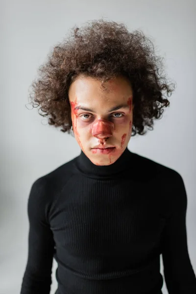 Раненый африканский американец со сломанным носом и кровоточащим лицом, смотрящий на камеру, изолированную на сером — стоковое фото