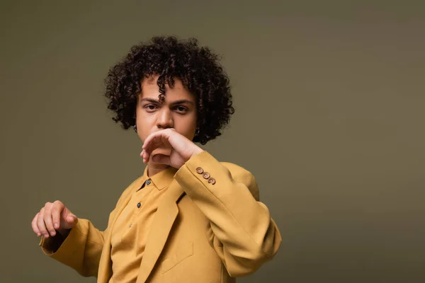 Jovem e moderno homem americano africano em amarelo casual traje segurando mão perto do rosto enquanto olha para a câmera isolada no cinza — Fotografia de Stock