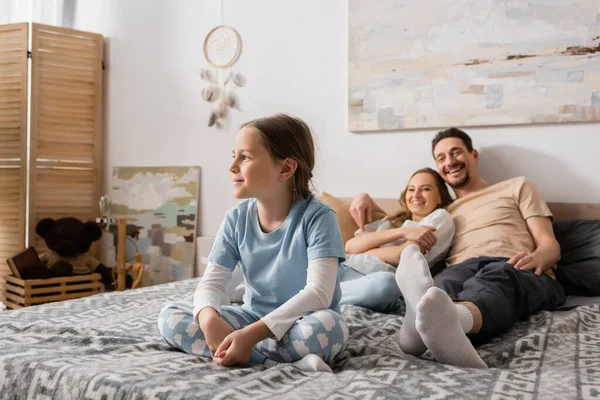 Cheerful girl sitting on bed and looking away near blurred parents resting on background — Fotografia de Stock