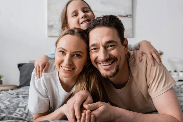 Portrait of happy child hugging cheerful parents looking at camera while resting in bedroom — Fotografia de Stock