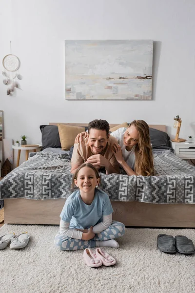 Веселый ребенок сидит на ковре вокруг тапочки около счастливых родителей на кровати — стоковое фото